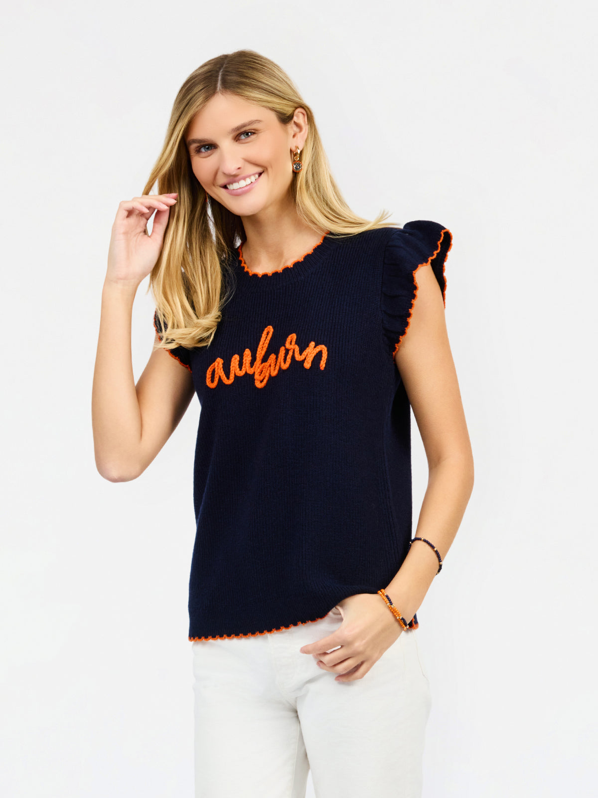 A woman modeling a navy blue ribbed knit top with orange trim and 'auburn' script across the chest, paired with white pants against a light gray studio background.
