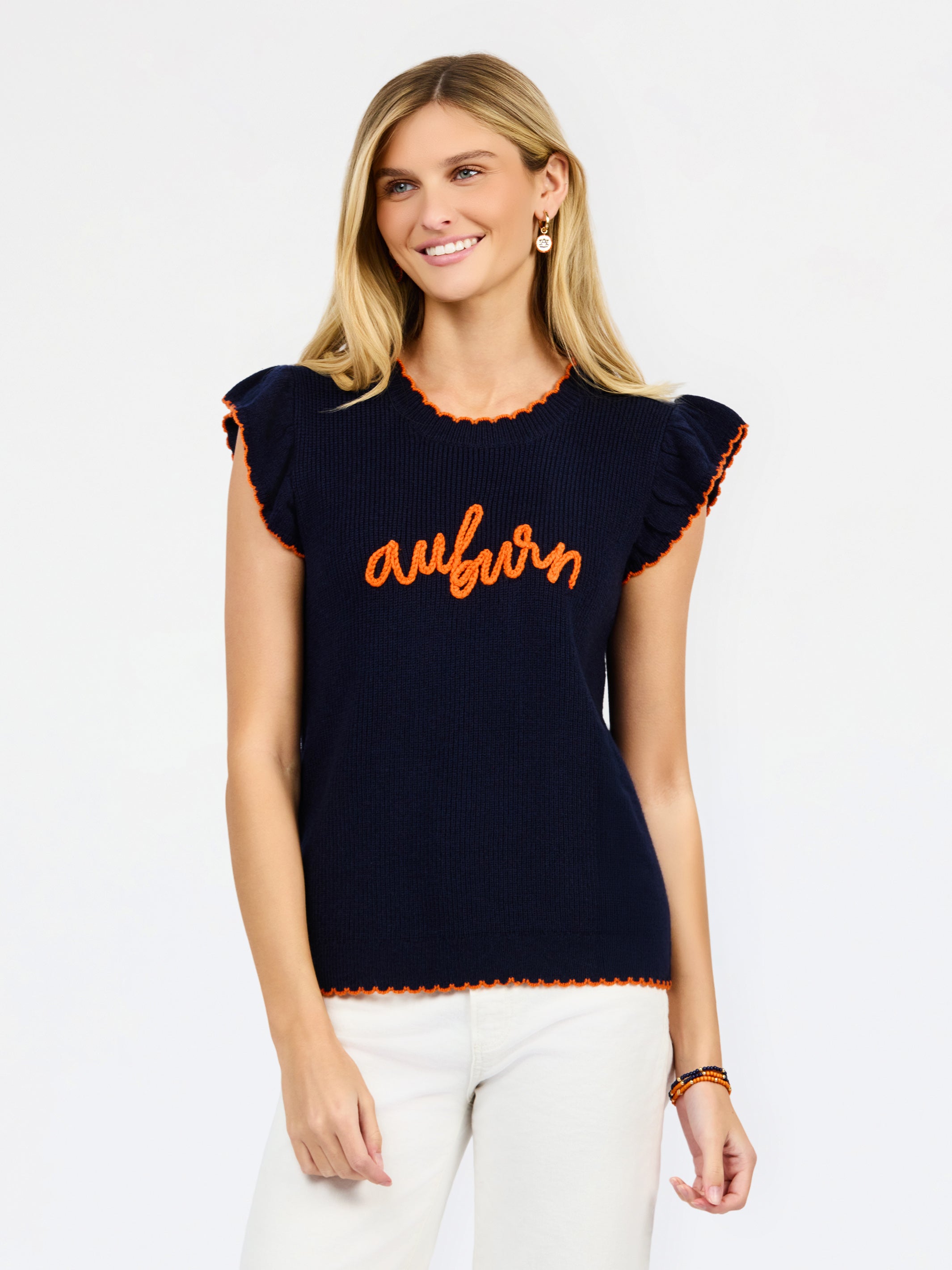 A woman modeling a navy blue ribbed knit top with orange trim and 'auburn' script across the chest, paired with white pants against a light gray studio background.