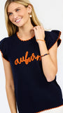 A woman modeling a navy blue ribbed knit top with orange trim and 'auburn' script across the chest, paired with white pants against a light gray studio background.