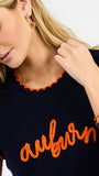 A woman modeling a navy blue ribbed knit top with orange trim and 'auburn' script across the chest, paired with white pants against a light gray studio background.