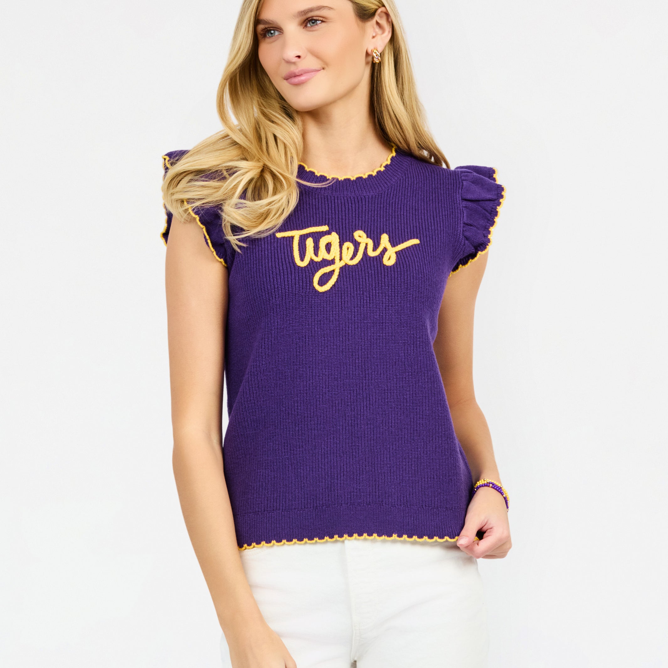 A woman modeling a purple ribbed knit top with yellow ruffle trim and 'Tigers' written across the chest in yellow script, paired with white pants against a light gray studio background.