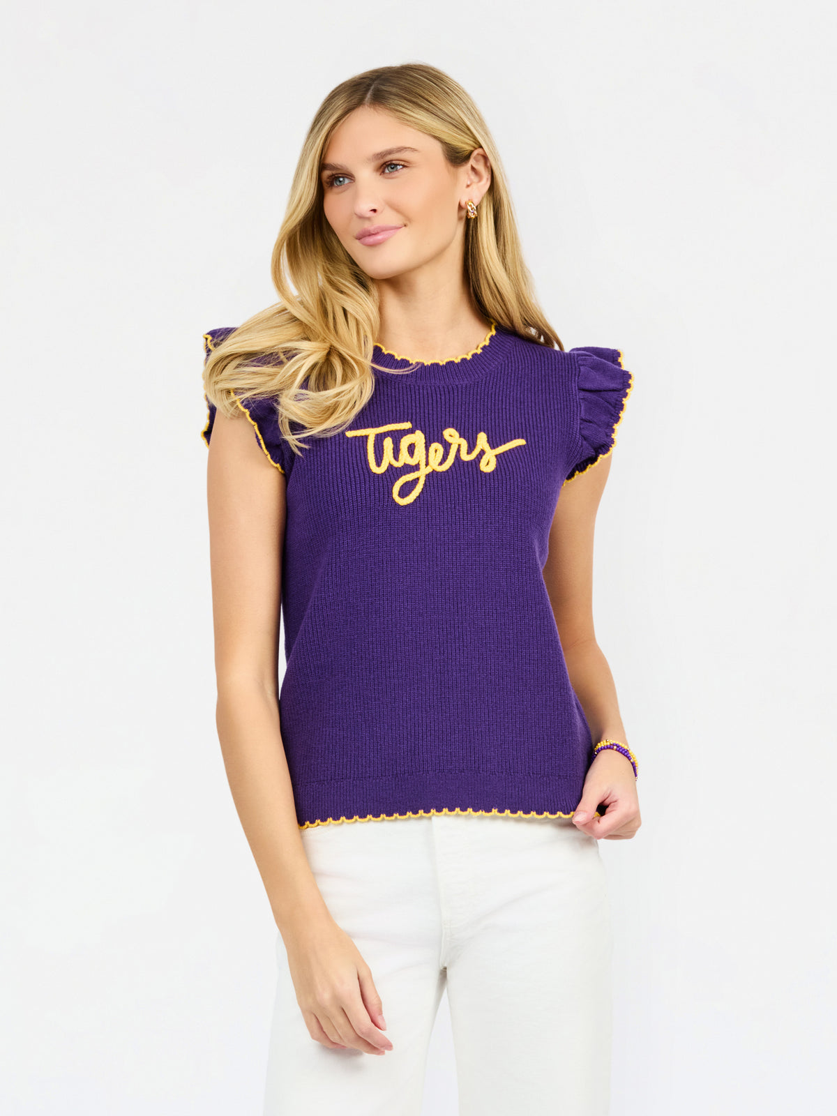 A woman modeling a purple ribbed knit top with yellow ruffle trim and 'Tigers' written across the chest in yellow script, paired with white pants against a light gray studio background.