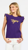 A woman modeling a purple ribbed knit top with yellow ruffle trim and 'Tigers' written across the chest in yellow script, paired with white pants against a light gray studio background.