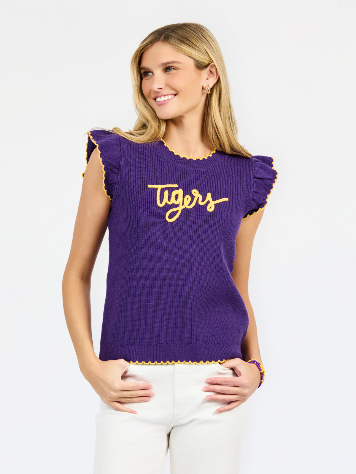 A woman modeling a purple ribbed knit top with yellow ruffle trim and 'Tigers' written across the chest in yellow script, paired with white pants against a light gray studio background.
