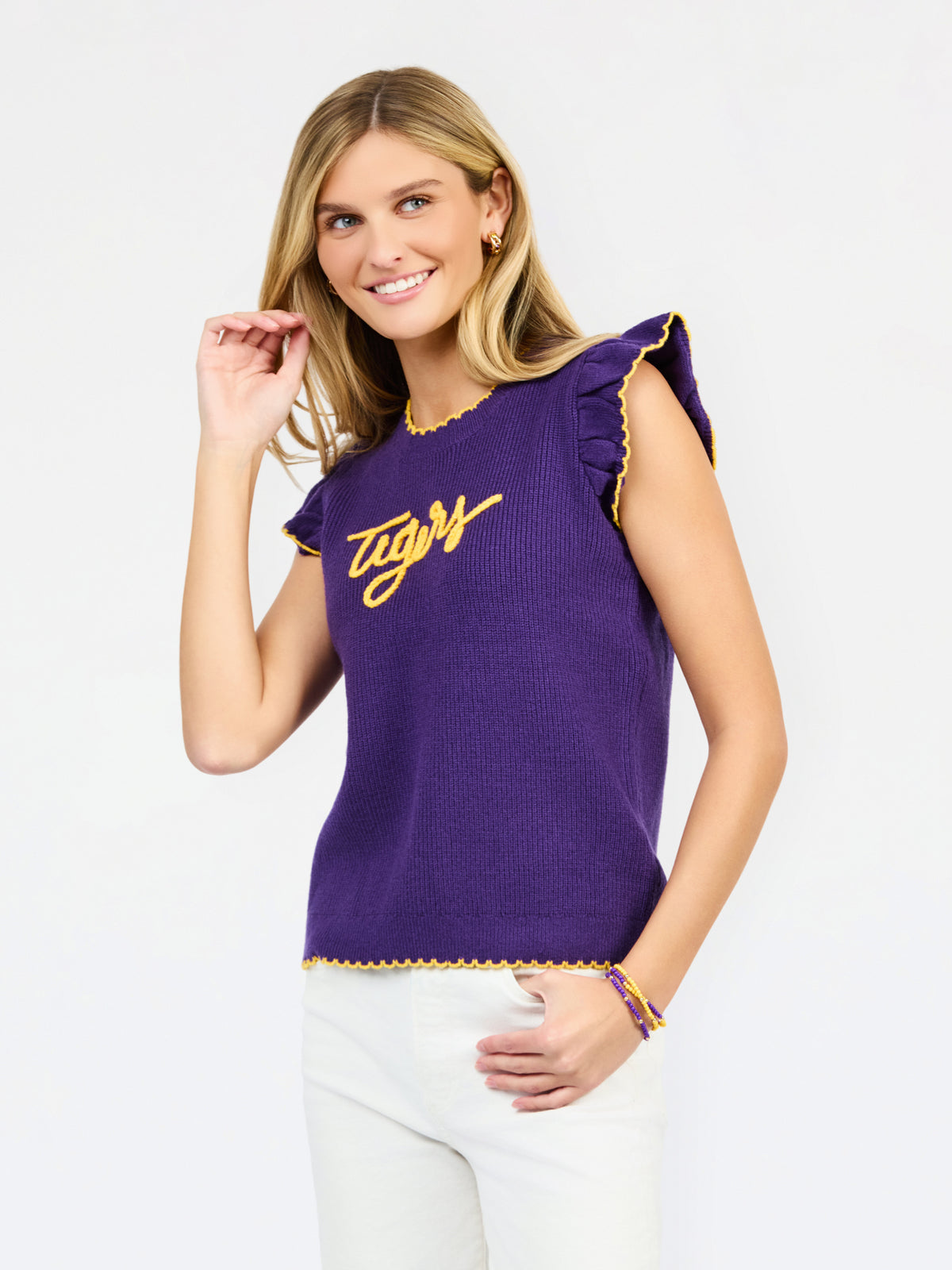A woman modeling a purple ribbed knit top with yellow ruffle trim and 'Tigers' written across the chest in yellow script, paired with white pants against a light gray studio background.