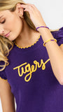 A woman modeling a purple ribbed knit top with yellow ruffle trim and 'Tigers' written across the chest in yellow script, paired with white pants against a light gray studio background.