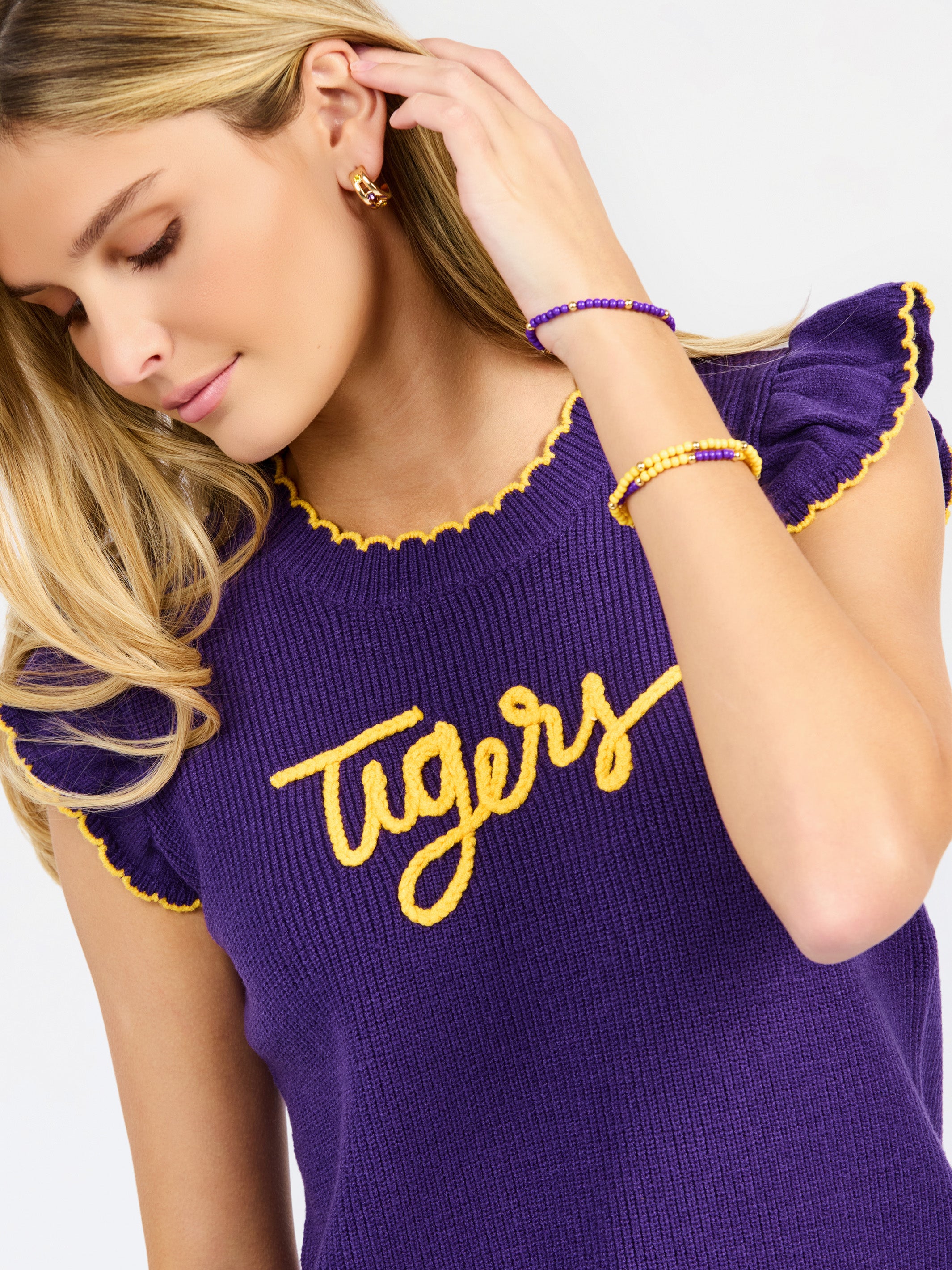 A woman modeling a purple ribbed knit top with yellow ruffle trim and 'Tigers' written across the chest in yellow script, paired with white pants against a light gray studio background.