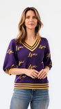A woman modeling a purple knit sweater with yellow and white striped trim and 'LSU' script lettering, paired with blue jeans against a plain white background.
