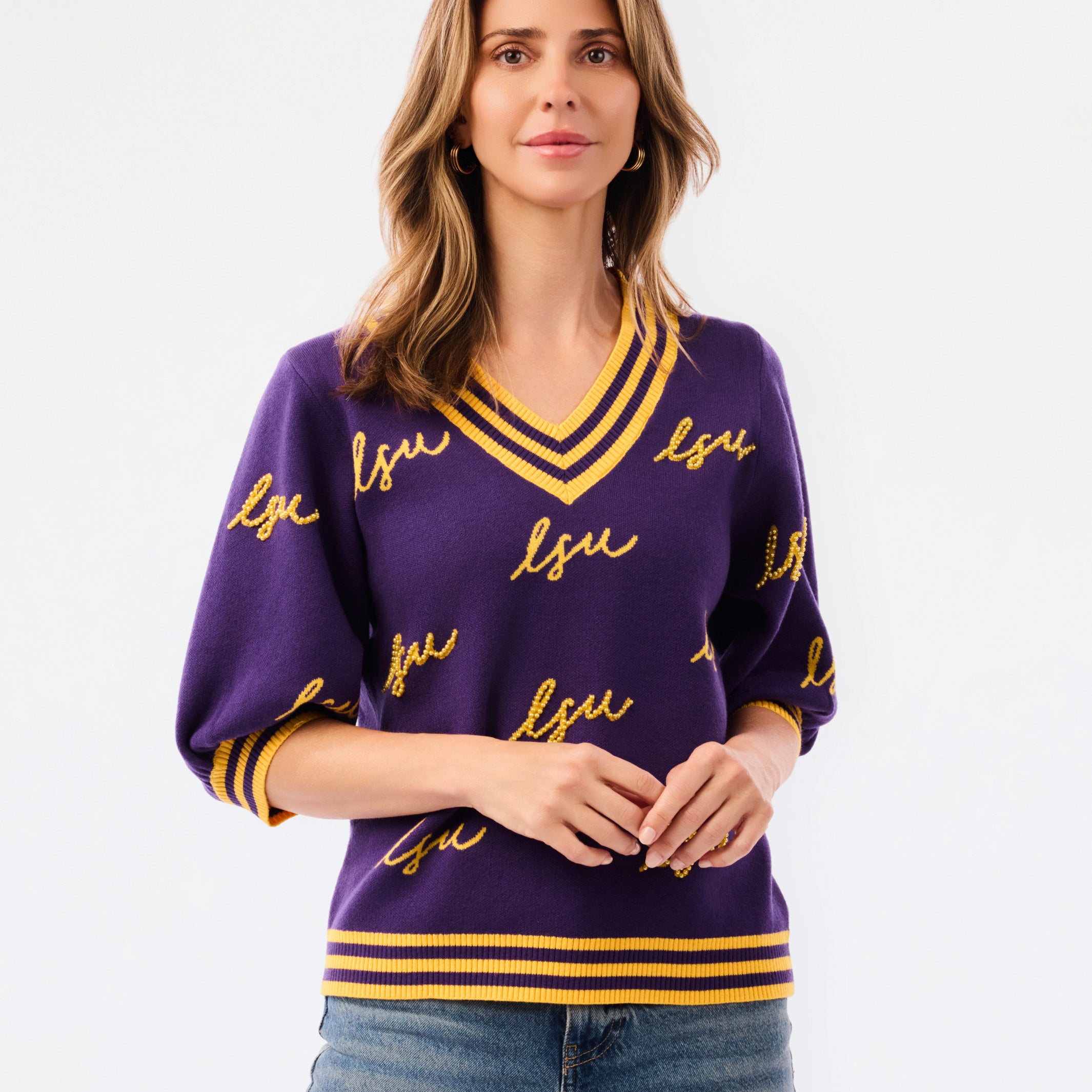 A woman modeling a purple knit sweater with yellow and white striped trim and 'LSU' script lettering, paired with blue jeans against a plain white background.