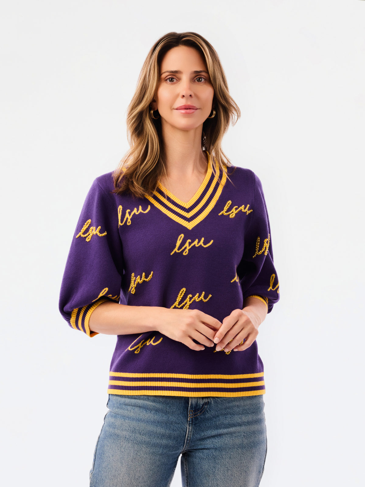 A woman modeling a purple knit sweater with yellow and white striped trim and 'LSU' script lettering, paired with blue jeans against a plain white background.