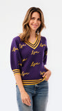A woman modeling a purple knit sweater with yellow and white striped trim and 'LSU' script lettering, paired with blue jeans against a plain white background.