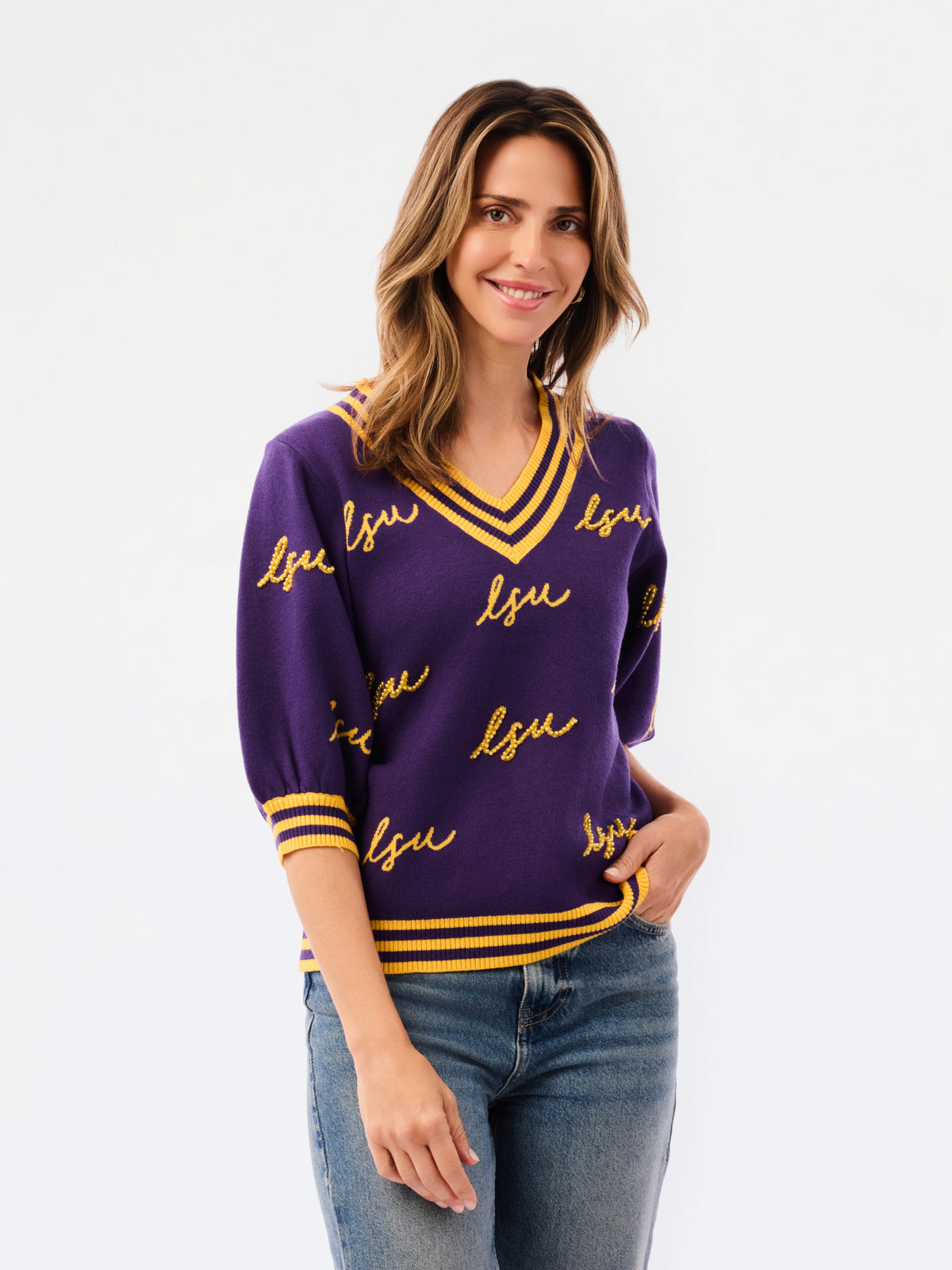 A woman modeling a purple knit sweater with yellow and white striped trim and 'LSU' script lettering, paired with blue jeans against a plain white background.
