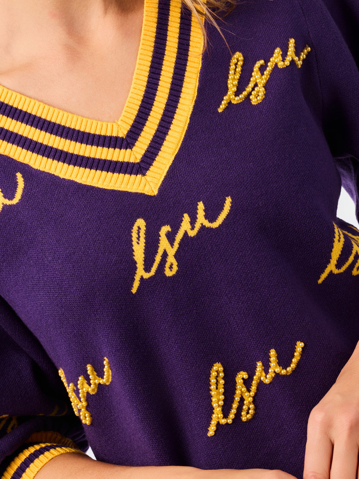 A woman modeling a purple knit sweater with yellow and white striped trim and 'LSU' script lettering, paired with blue jeans against a plain white background.