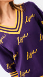 A woman modeling a purple knit sweater with yellow and white striped trim and 'LSU' script lettering, paired with blue jeans against a plain white background.