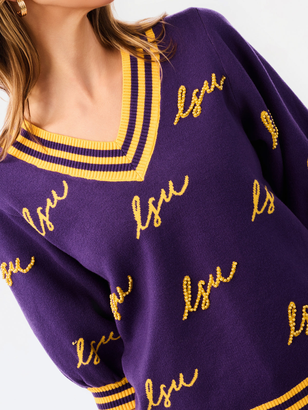 A woman modeling a purple knit sweater with yellow and white striped trim and 'LSU' script lettering, paired with blue jeans against a plain white background.
