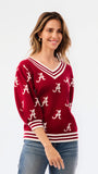 A woman wearing a crimson red V-neck sweater with white stripes and white Alabama 'A' logos, paired with blue jeans, against a plain white background.