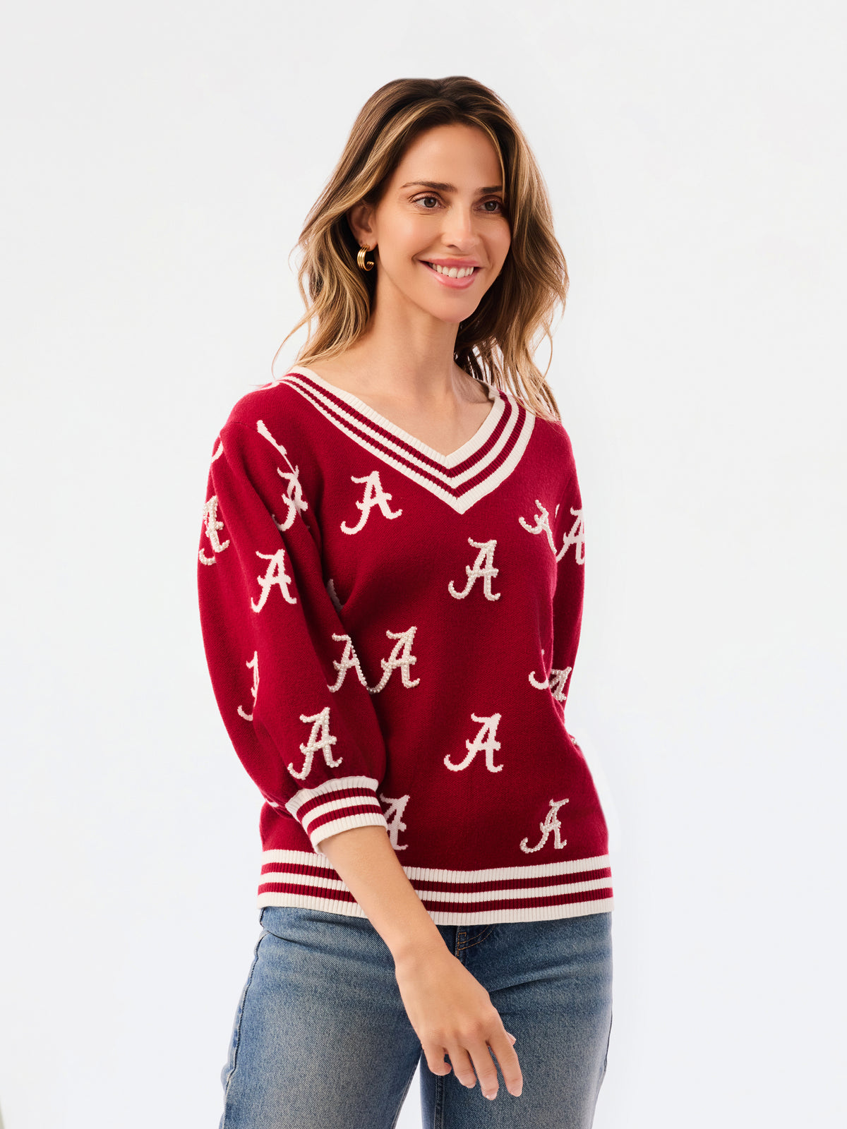 A woman wearing a crimson red V-neck sweater with white stripes and white Alabama 'A' logos, paired with blue jeans, against a plain white background.