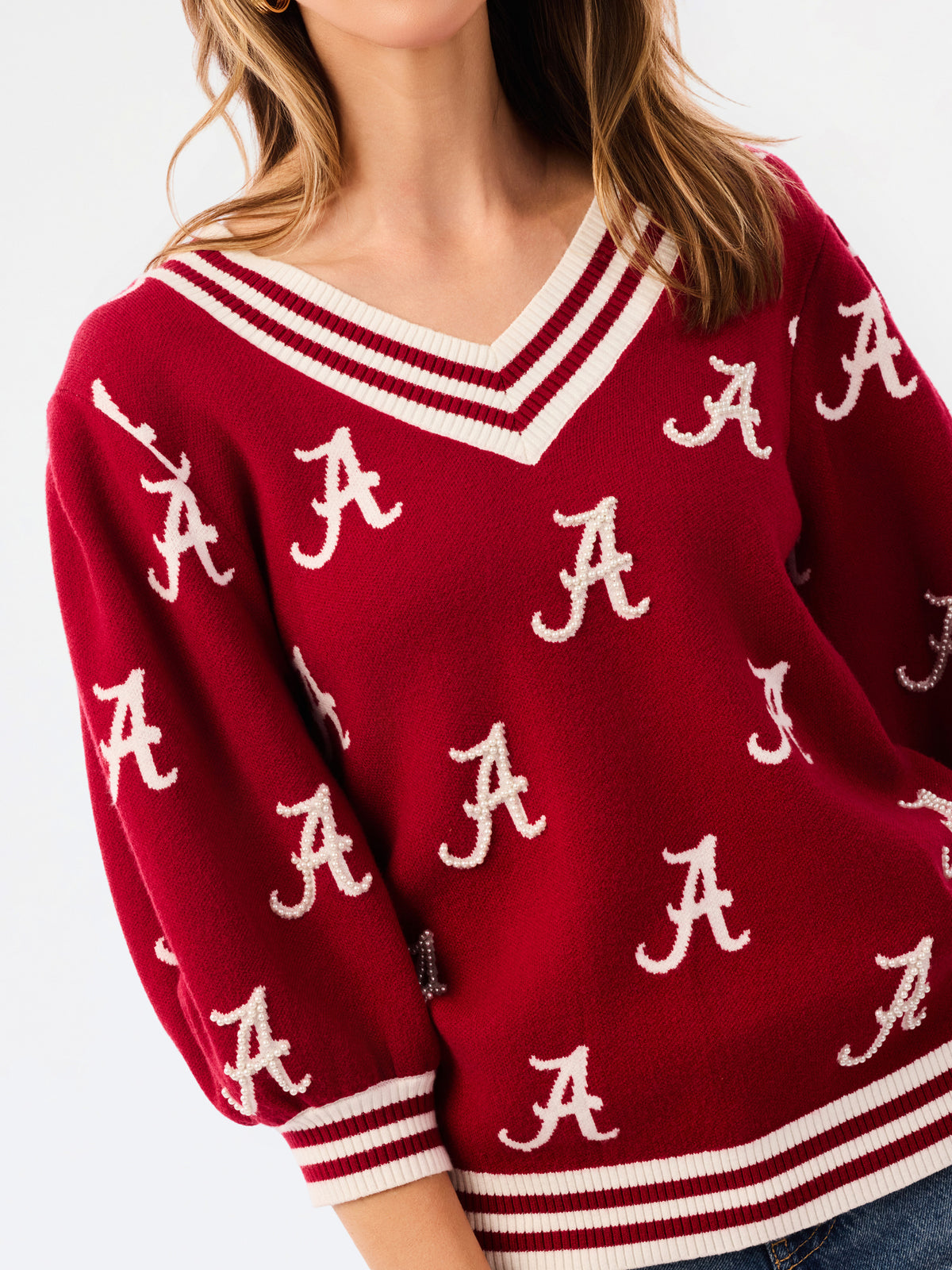 A woman wearing a crimson red V-neck sweater with white stripes and white Alabama 'A' logos, paired with blue jeans, against a plain white background.