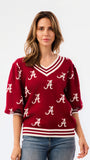 A woman wearing a crimson red V-neck sweater with white stripes and white Alabama 'A' logos, paired with blue jeans, against a plain white background.