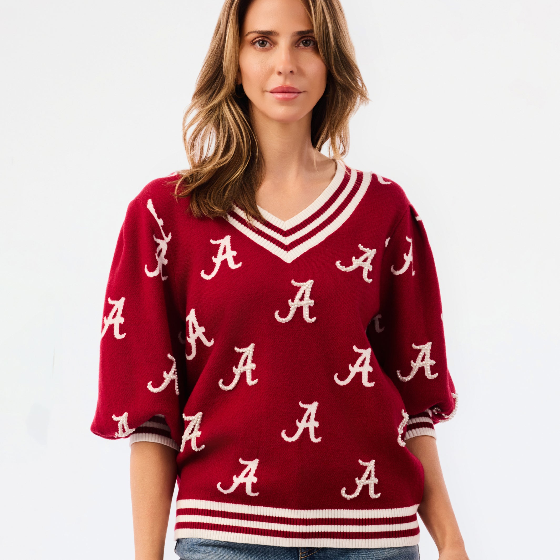 A woman wearing a crimson red V-neck sweater with white stripes and white Alabama 'A' logos, paired with blue jeans, against a plain white background.