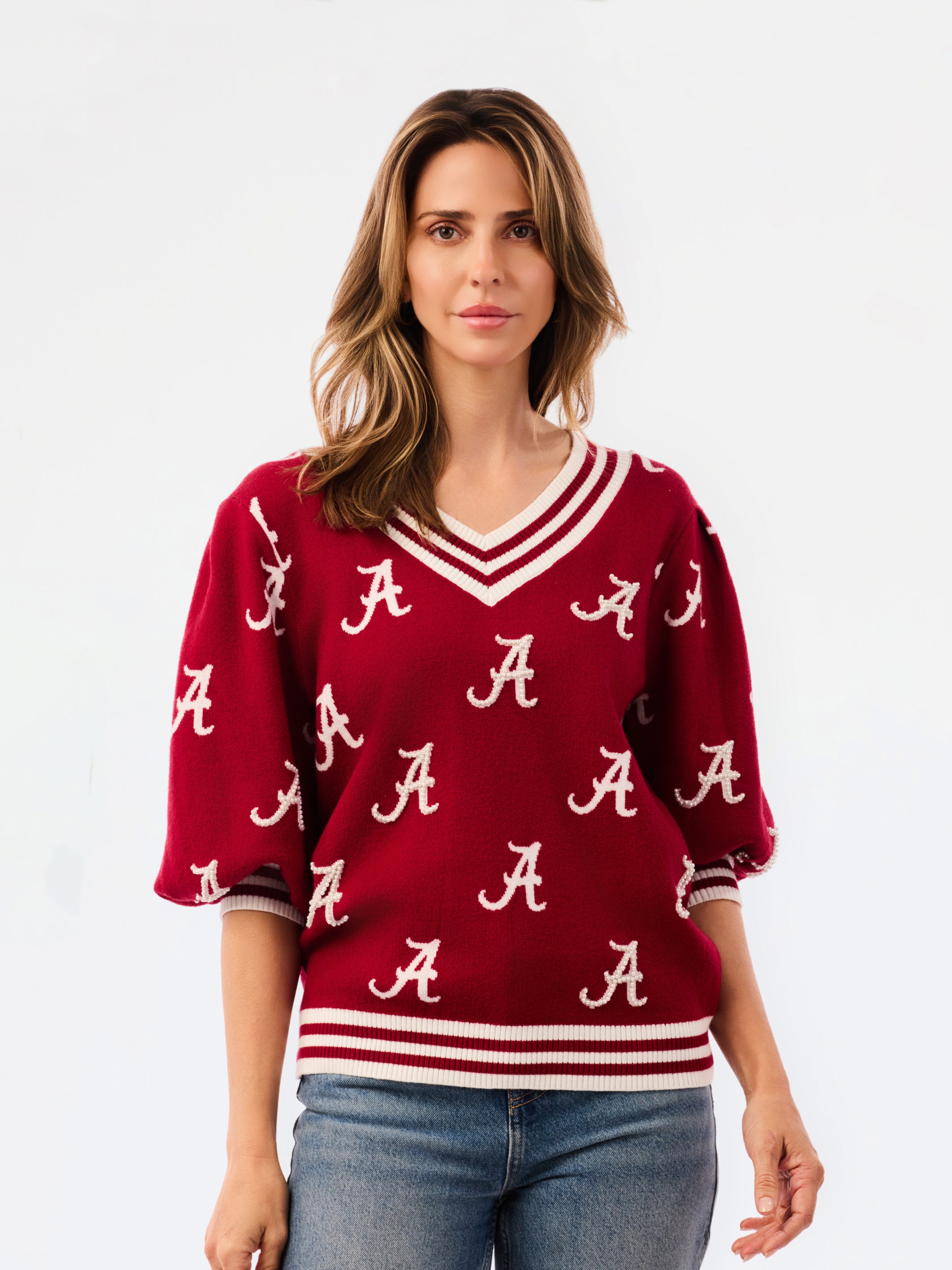 A woman wearing a crimson red V-neck sweater with white stripes and white Alabama 'A' logos, paired with blue jeans, against a plain white background.