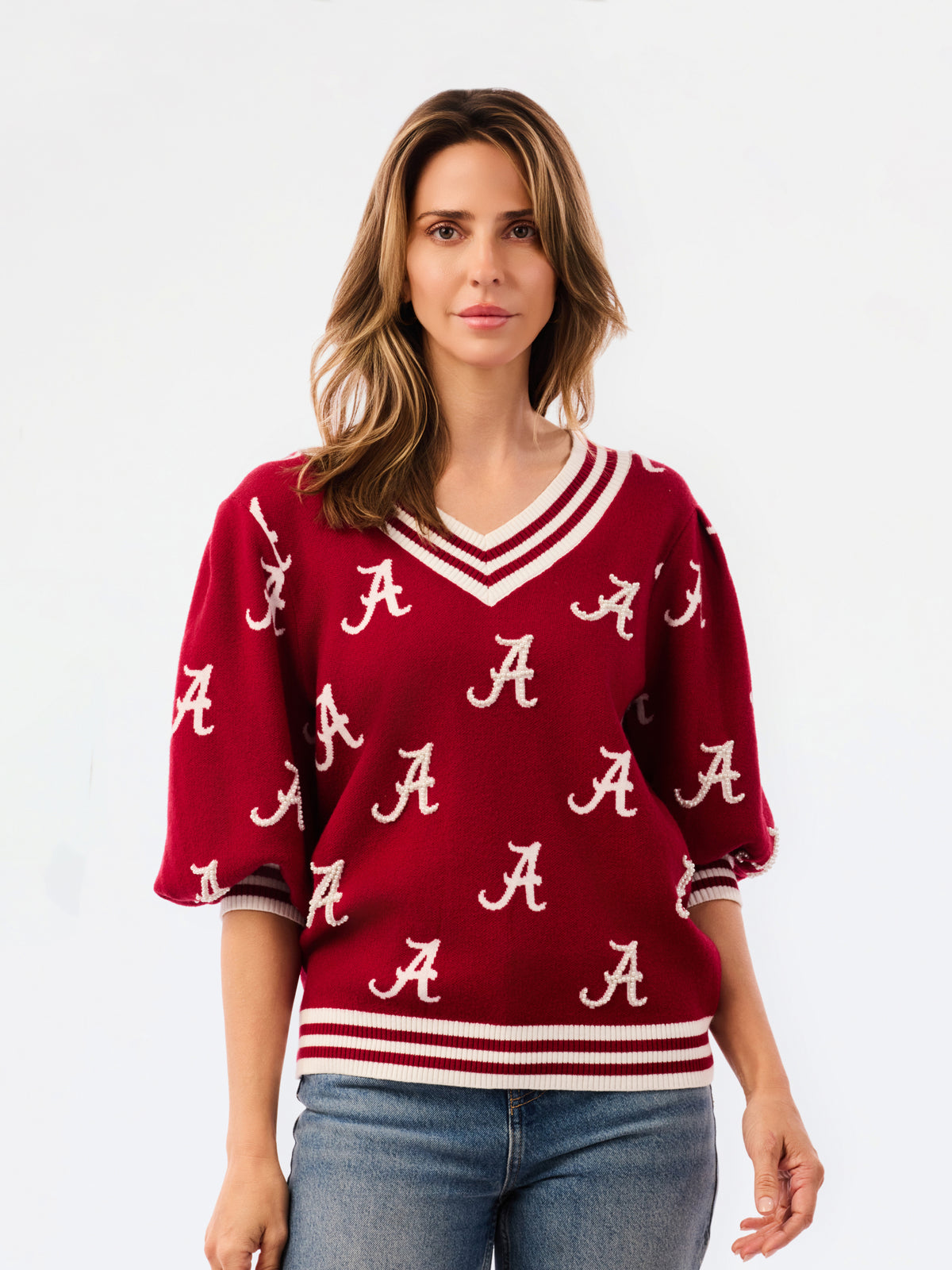 A woman wearing a crimson red V-neck sweater with white stripes and white Alabama 'A' logos, paired with blue jeans, against a plain white background.