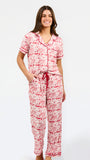 A woman modeling a two-piece pajama set with a red and white toile-style print, featuring a short-sleeve button-front top with a red tie collar and matching drawstring pants, against a plain white studio background.