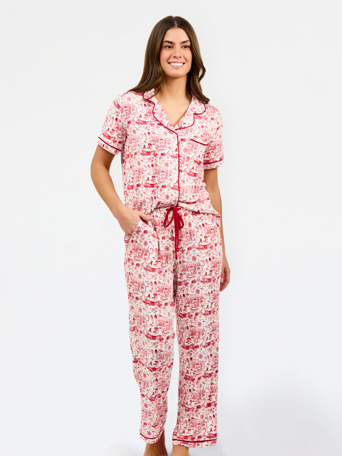 A woman modeling a two-piece pajama set with a red and white toile-style print, featuring a short-sleeve button-front top with a red tie collar and matching drawstring pants, against a plain white studio background.