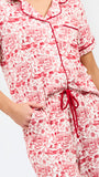 A woman modeling a two-piece pajama set with a red and white toile-style print, featuring a short-sleeve button-front top with a red tie collar and matching drawstring pants, against a plain white studio background.