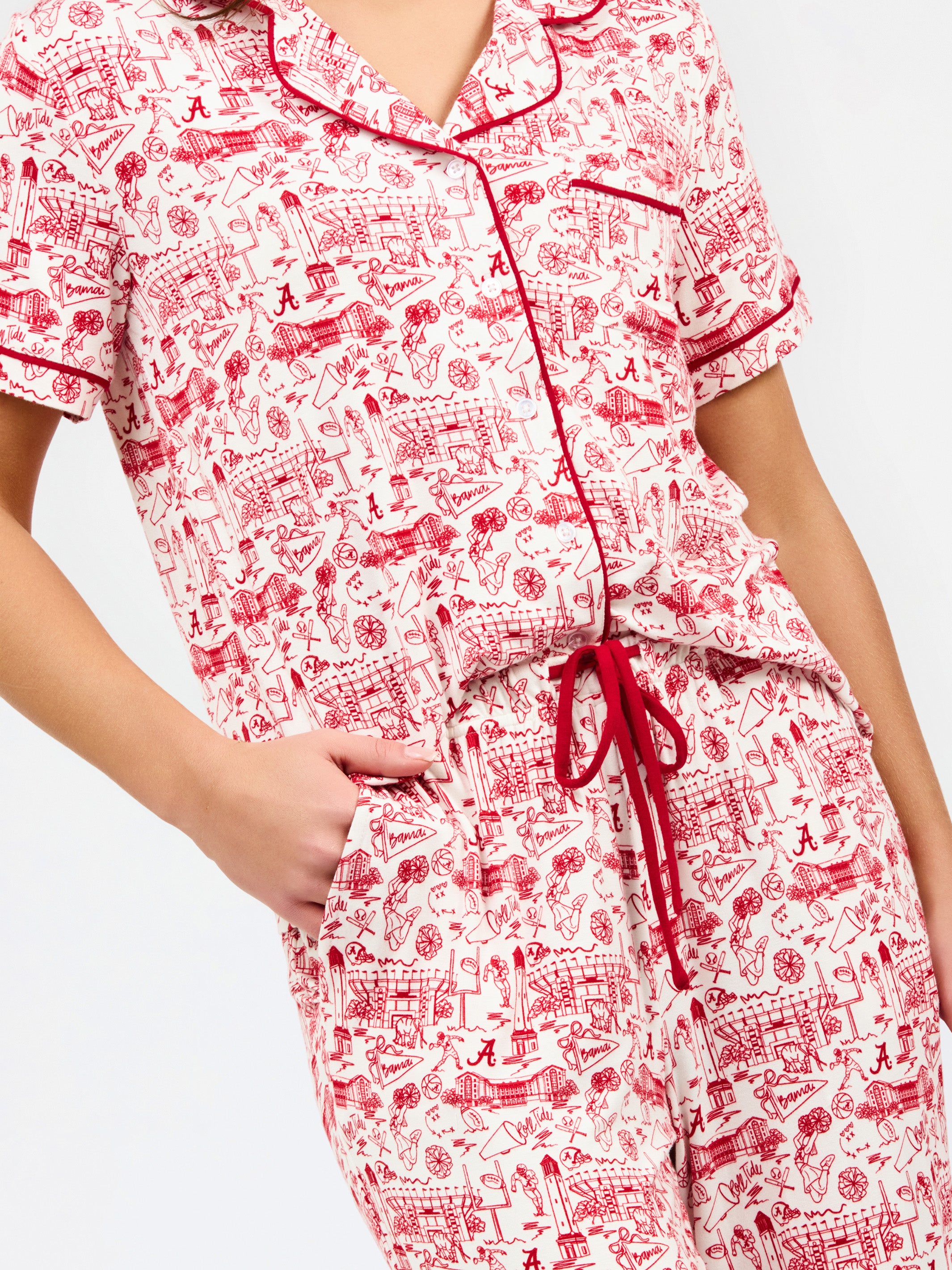 A woman modeling a two-piece pajama set with a red and white toile-style print, featuring a short-sleeve button-front top with a red tie collar and matching drawstring pants, against a plain white studio background.