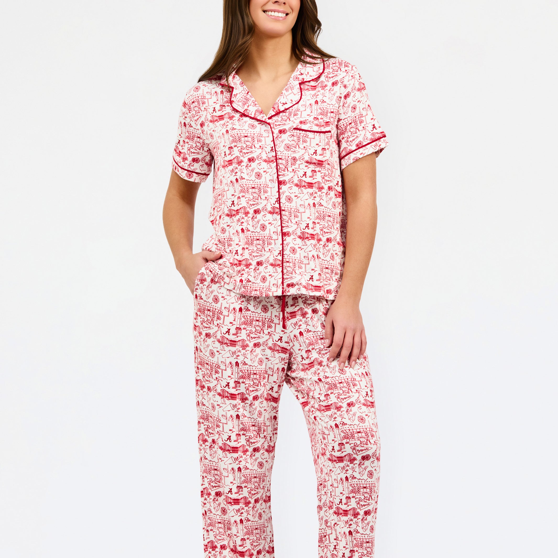A woman modeling a two-piece pajama set with a red and white toile-style print, featuring a short-sleeve button-front top with a red tie collar and matching drawstring pants, against a plain white studio background.