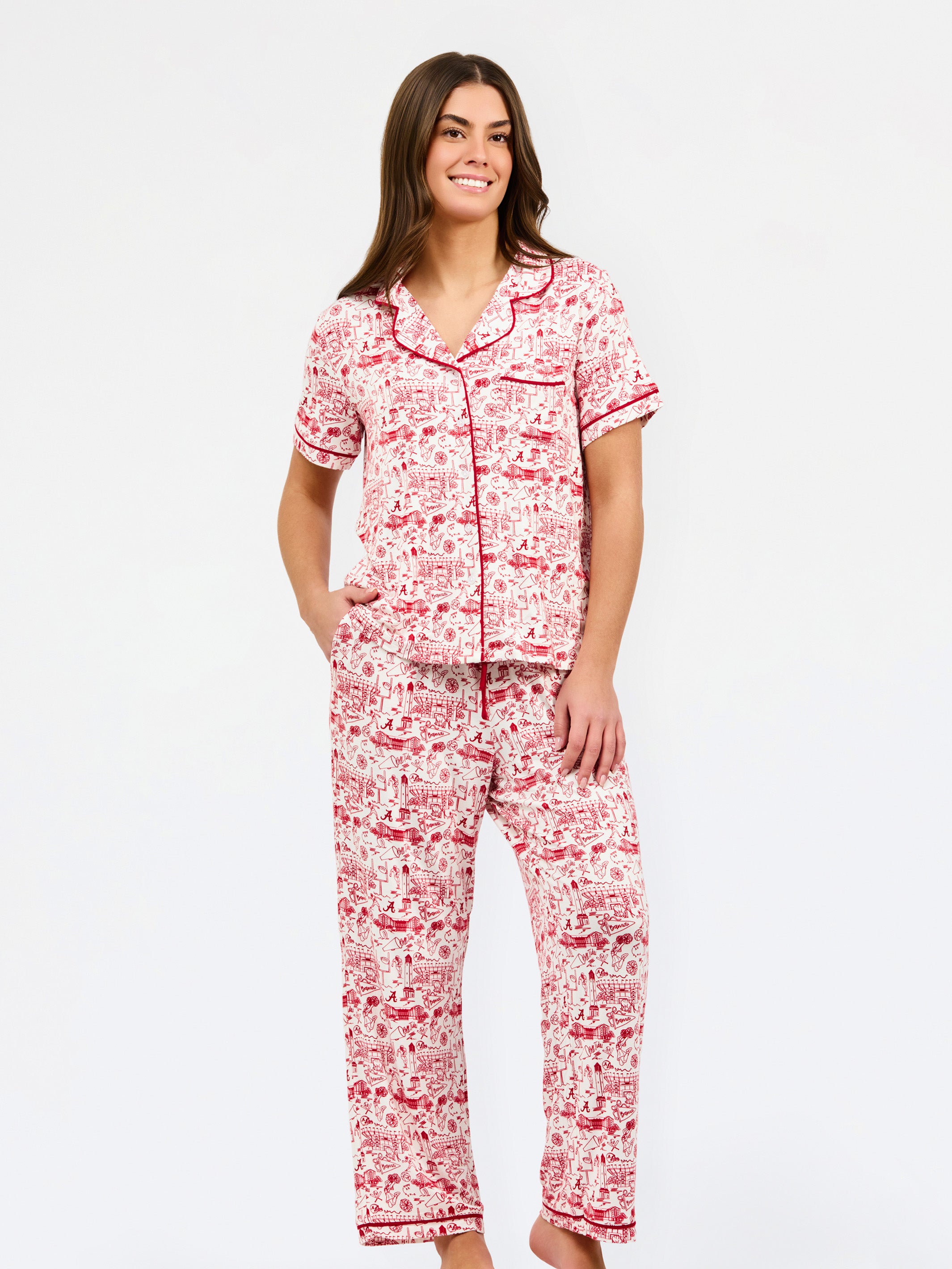 A woman modeling a two-piece pajama set with a red and white toile-style print, featuring a short-sleeve button-front top with a red tie collar and matching drawstring pants, against a plain white studio background.