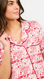 A woman modeling a two-piece pajama set with a red and white toile-style print, featuring a short-sleeve button-front top with a red tie collar and matching drawstring pants, against a plain white studio background.