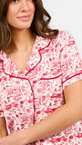 A woman modeling a two-piece pajama set with a red and white toile-style print, featuring a short-sleeve button-front top with a red tie collar and matching drawstring pants, against a plain white studio background.