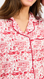 A woman modeling a two-piece pajama set with a red and white toile-style print, featuring a short-sleeve button-front top with a red tie collar and matching drawstring pants, against a plain white studio background.