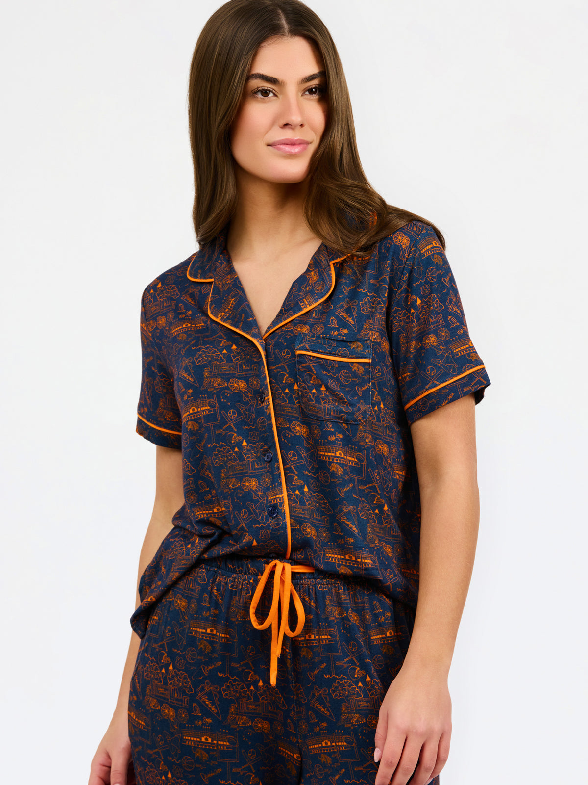 A woman modeling a short-sleeve pajama set with an orange and navy blue patterned design, featuring a collared top with a front zipper and drawstring, and matching pants.