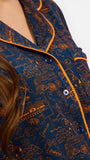 A woman modeling a short-sleeve pajama set with an orange and navy blue patterned design, featuring a collared top with a front zipper and drawstring, and matching pants.