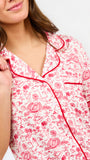 A woman modeling a two-piece pajama set featuring a short-sleeve button-front top and matching pants, red toile print with red piping and a red drawstring waist.