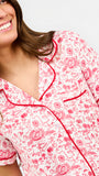 A woman modeling a two-piece pajama set featuring a short-sleeve button-front top and matching pants, red toile print with red piping and a red drawstring waist.