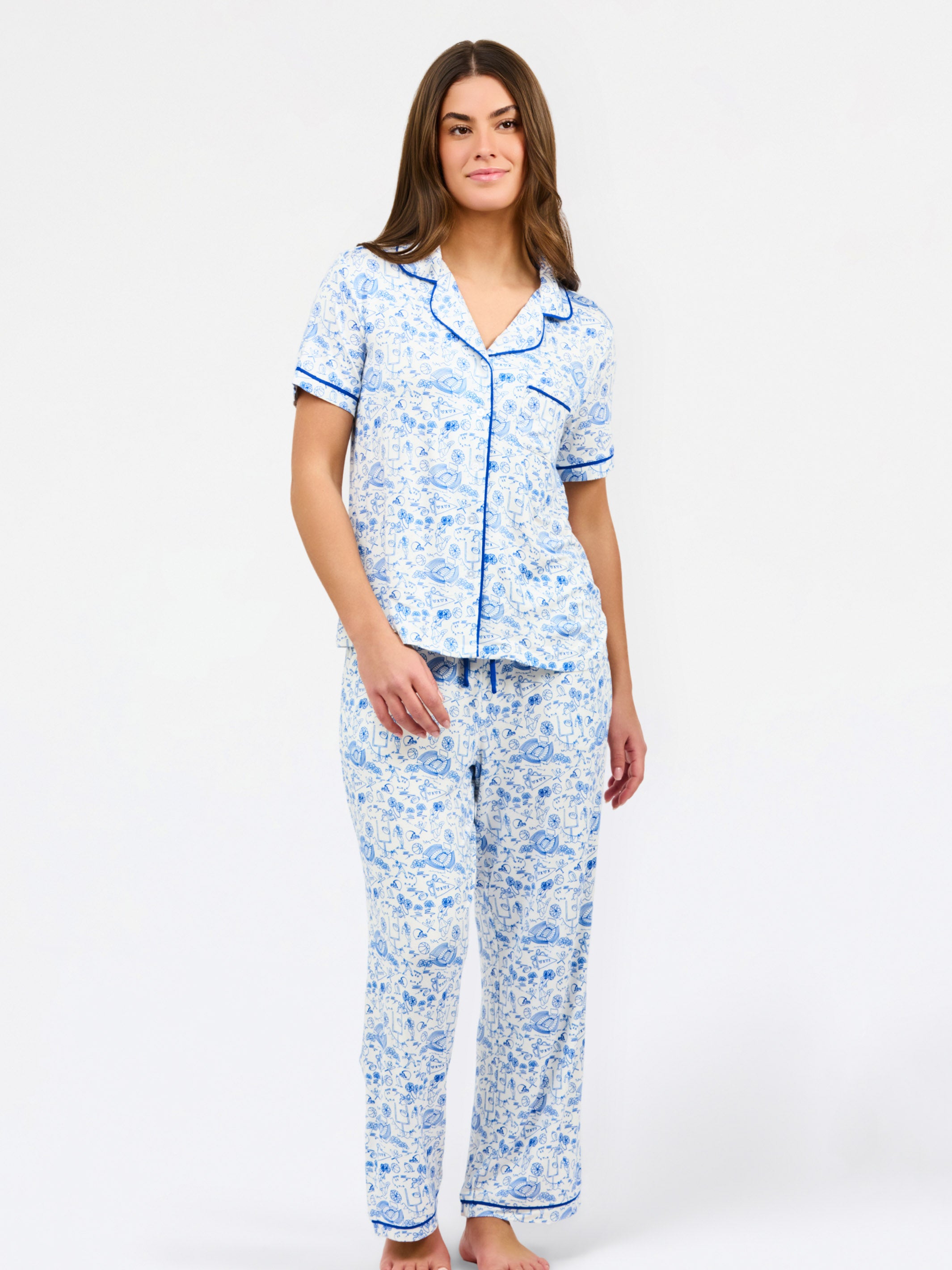 A woman modeling a two-piece pajama set featuring a short-sleeve button-up top and matching pants in a blue and white toile pattern, set against a plain white studio background.