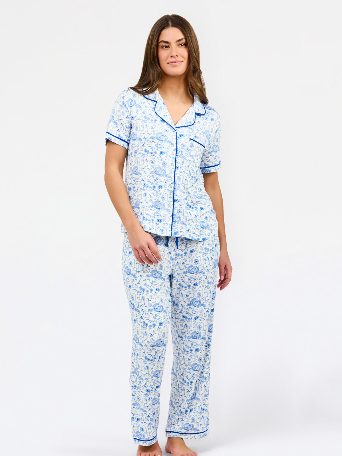 A woman modeling a two-piece pajama set featuring a short-sleeve button-up top and matching pants in a blue and white toile pattern, set against a plain white studio background.