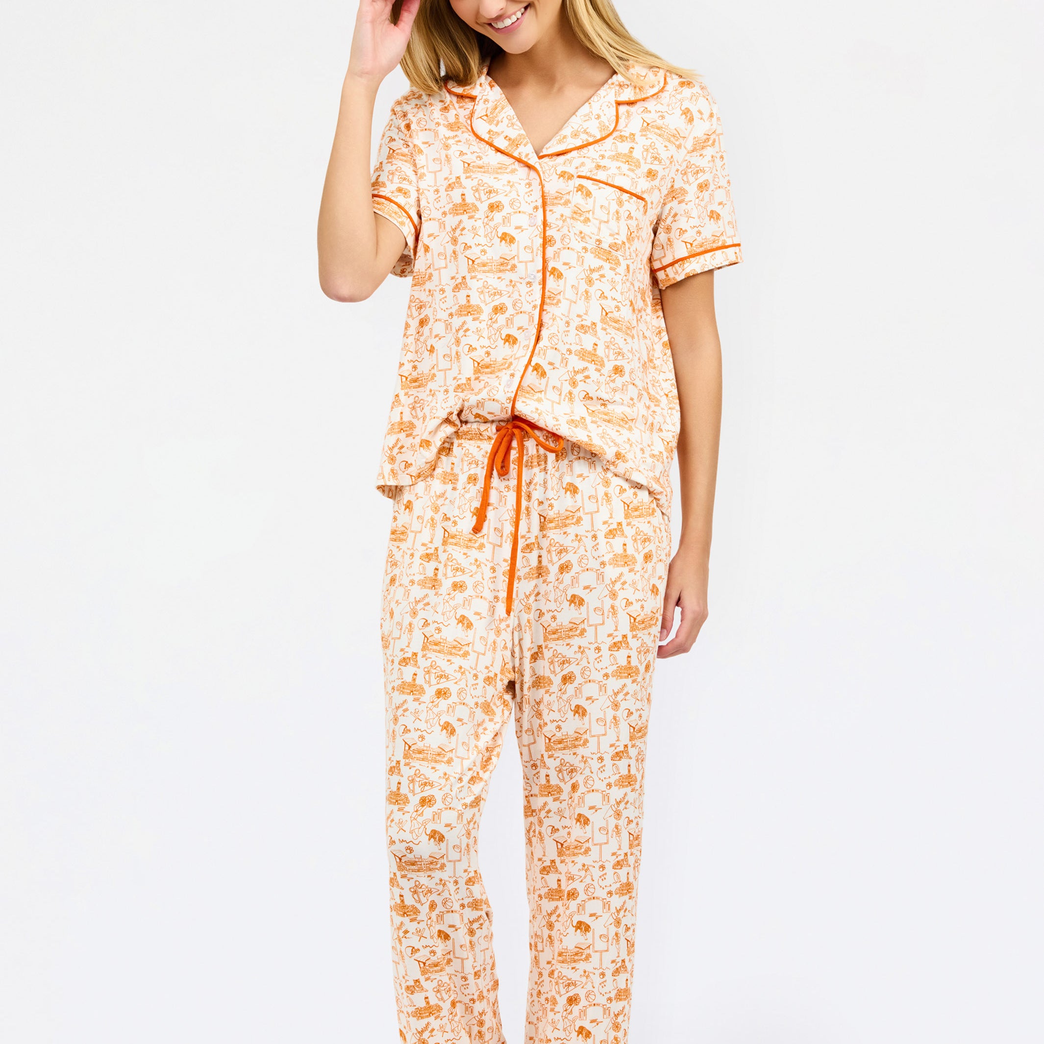 A woman modeling a two-piece short-sleeve pajama set with an orange and cream floral print, featuring a button-up top and matching pants with a drawstring waist.