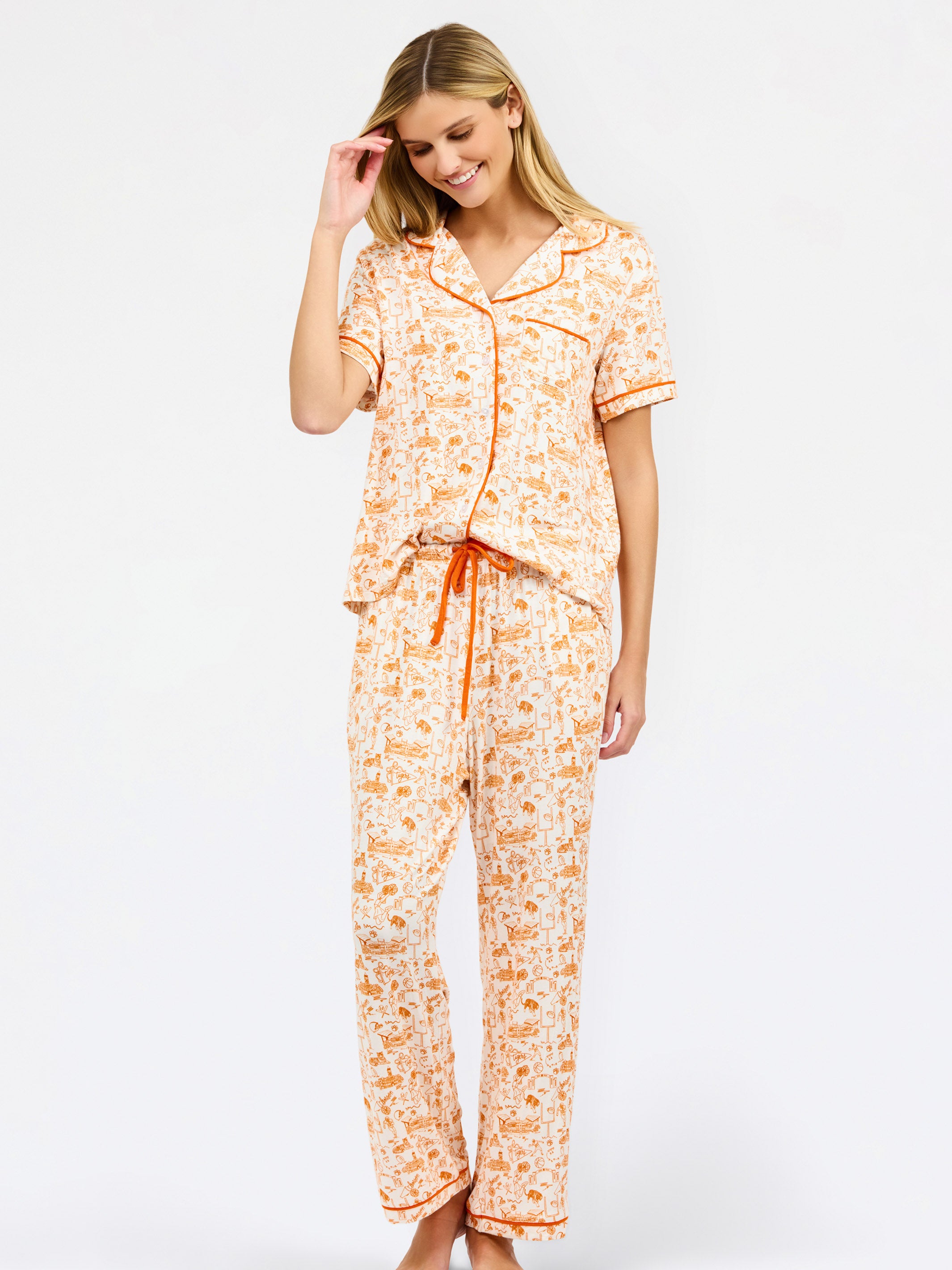 A woman modeling a two-piece short-sleeve pajama set with an orange and cream floral print, featuring a button-up top and matching pants with a drawstring waist.