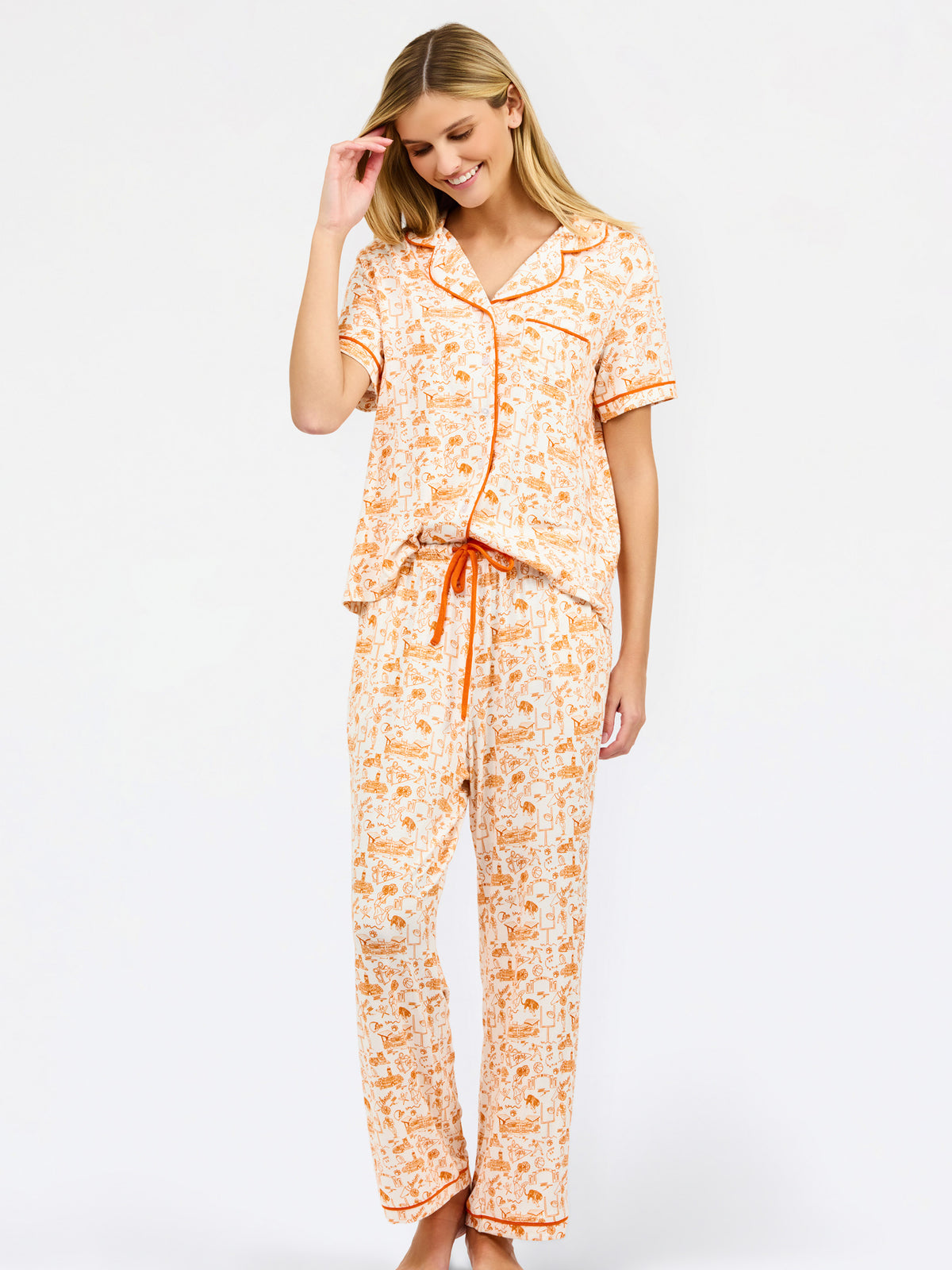 A woman modeling a two-piece short-sleeve pajama set with an orange and cream floral print, featuring a button-up top and matching pants with a drawstring waist.