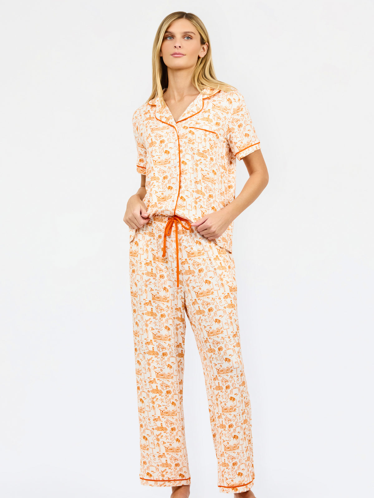 A woman modeling a short-sleeve pajama set with an orange and cream floral print, featuring a button-up front, matching shorts, and a drawstring waist, set against a plain white studio background.