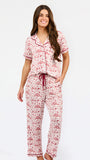 A woman modeling a two-piece pajama set with a short-sleeve button-front top and matching pants, featuring a pink and red floral pattern on a white background with dark red piping and a drawstring waist.