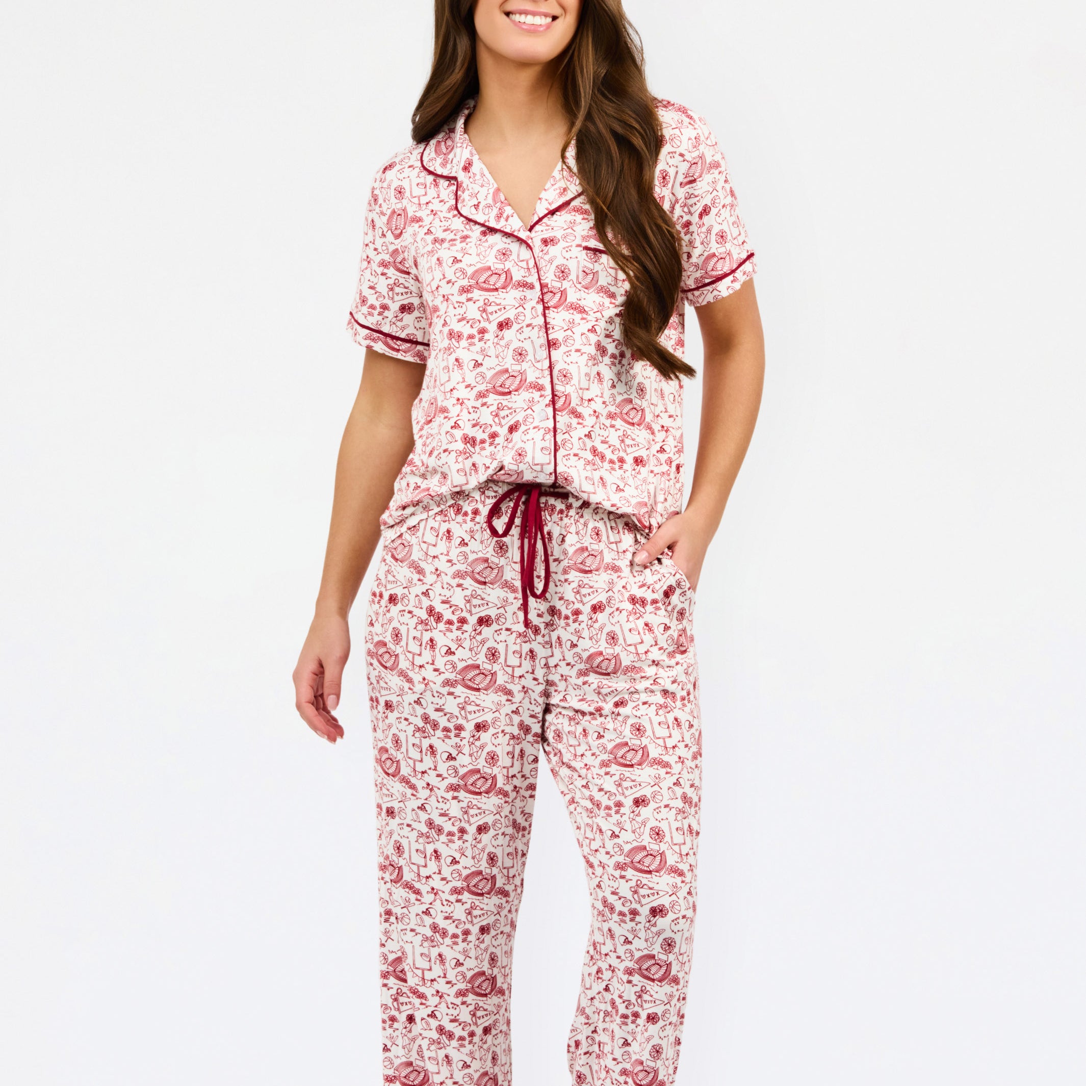 A woman modeling a two-piece pajama set with a short-sleeve button-front top and matching pants, featuring a pink and red floral pattern on a white background with dark red piping and a drawstring waist.