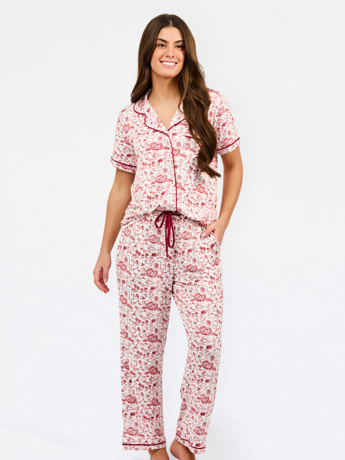 A woman modeling a two-piece pajama set with a short-sleeve button-front top and matching pants, featuring a pink and red floral pattern on a white background with dark red piping and a drawstring waist.