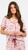 A woman modeling a two-piece pajama set with a short-sleeve button-front top and matching pants, featuring a pink and red floral pattern on a white background with dark red piping and a drawstring waist.