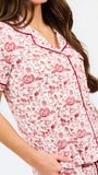 A woman modeling a two-piece pajama set with a short-sleeve button-front top and matching pants, featuring a pink and red floral pattern on a white background with dark red piping and a drawstring waist.